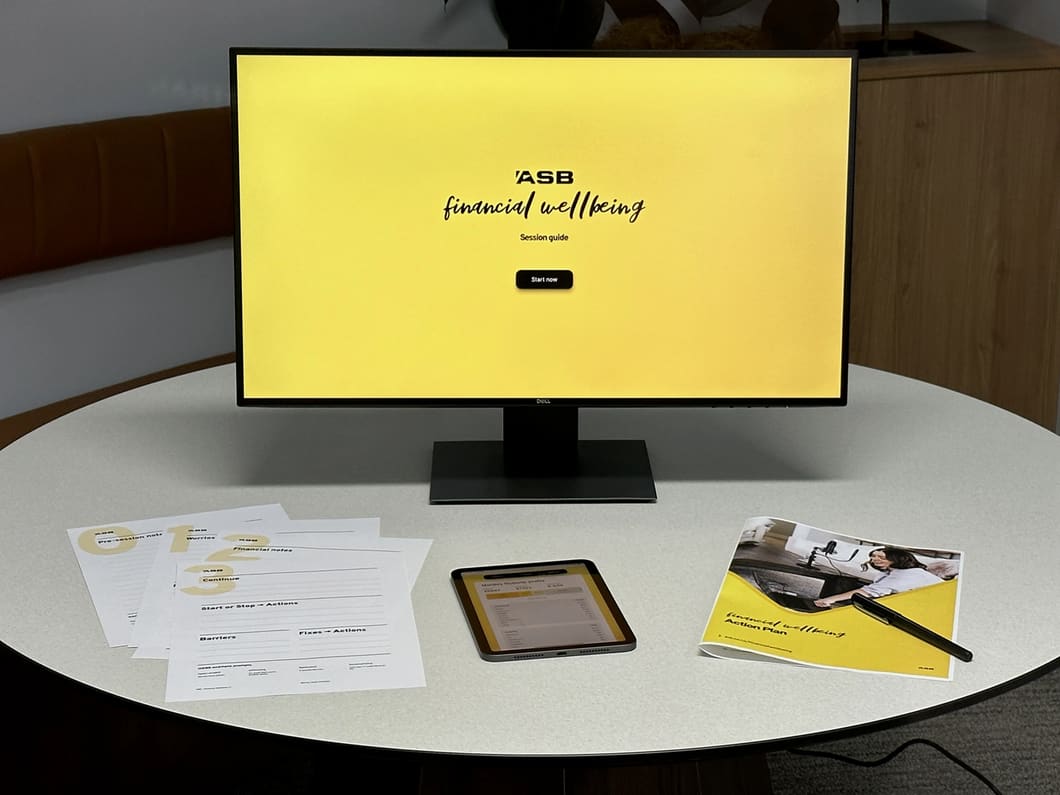 Ein Tisch mit einem Bildschirm, einem iPad und einem gedruckten Heft mit Arbeitsblättern — alles im Financial-Wellbeing-Branding.