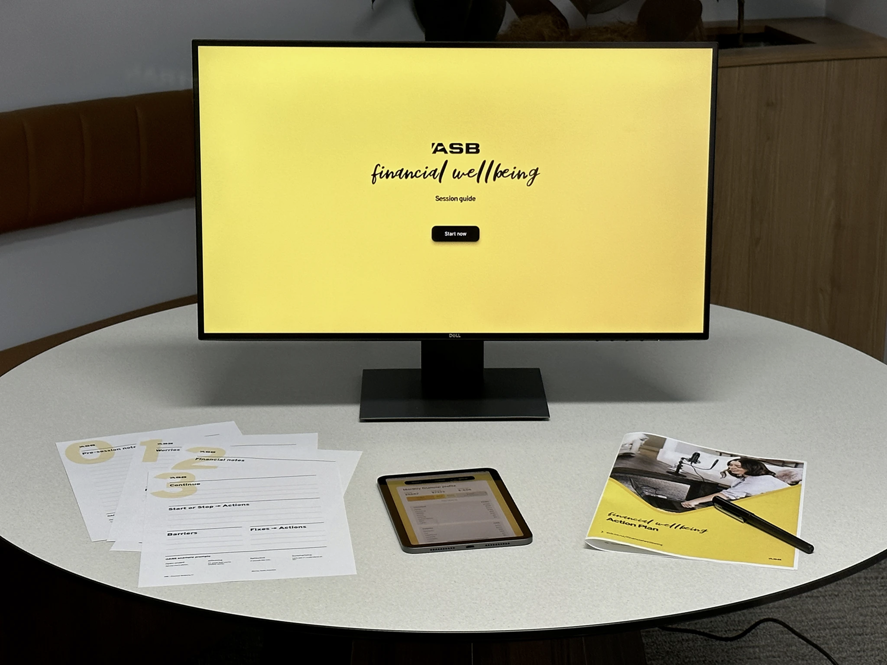 Ein Tisch mit einem Bildschirm, einem iPad und einem gedruckten Heft mit Arbeitsblättern — alles im Financial-Wellbeing-Branding.