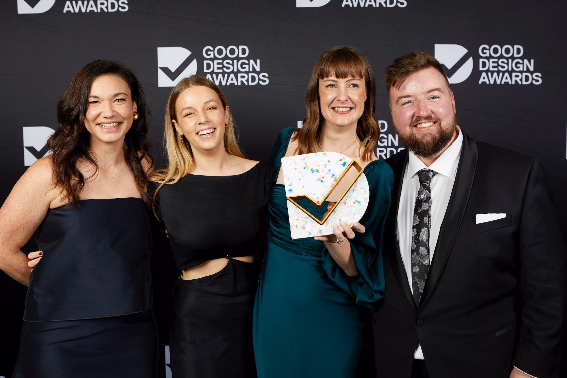 Drei Frauen und ein Mann stehen lächelnd nebeneinander vor einer Wand mit „Good Design Awards“-Logos. Sie sind für eine Black-Tie-Veranstaltung gekleidet, und eine der Frauen hält einen Award nach vorne.